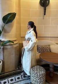 Lys beige skinn trenchcoat med vide ermer, parret med en lys tan håndveske. Står i et rom med dekorative planter og møbler.