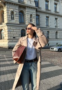 Μπεζ παλτό με ζώνη, λευκό τισερτάκι, ανοιχτό μπλε τζιν και καφέ δερμάτινη τσάντα. Γυαλιά ηλίου συμπληρώνουν το ντύσιμο. Πλακόστρωτος δρόμος και κτήρια στο βάθος.