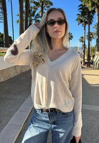 Pull en V couleur crème à manches longues, avec un patch marron sur le bras gauche. Porté avec un jean en denim bleu clair et une ceinture noire.