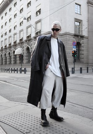 Persona con cabello corto y claro que lleva gafas de sol negras, abrigo largo de cuero negro, camisa blanca, pantalones claros y botas negras, de pie en una calle de la ciudad.