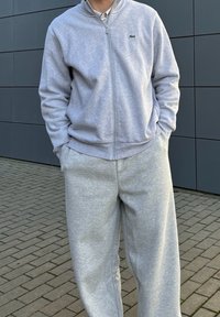 Γκρίζα ζακέτα με φερμουάρ και ταιριαστά παντελόνια από μαλακό ύφασμα. Η ζακέτα έχει ψηλό γιακά και ένα μικρό λογότυπο στην αριστερή πλευρά του στήθους. Χαλαρή εφαρμογή.