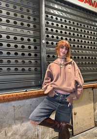 Brauner gekürzter Hoodie mit Kordelzügen, kombiniert mit verwaschenen schwarzen Denim-Shorts und kniehohen braunen Stiefeln mit Absatz, vor einem gemusterten Hintergrund.