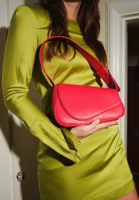 Bolso de cuero rojo con solapa curva y correa ajustable, sostenido contra un vestido de satén verde. Textura suave, diseño compacto.