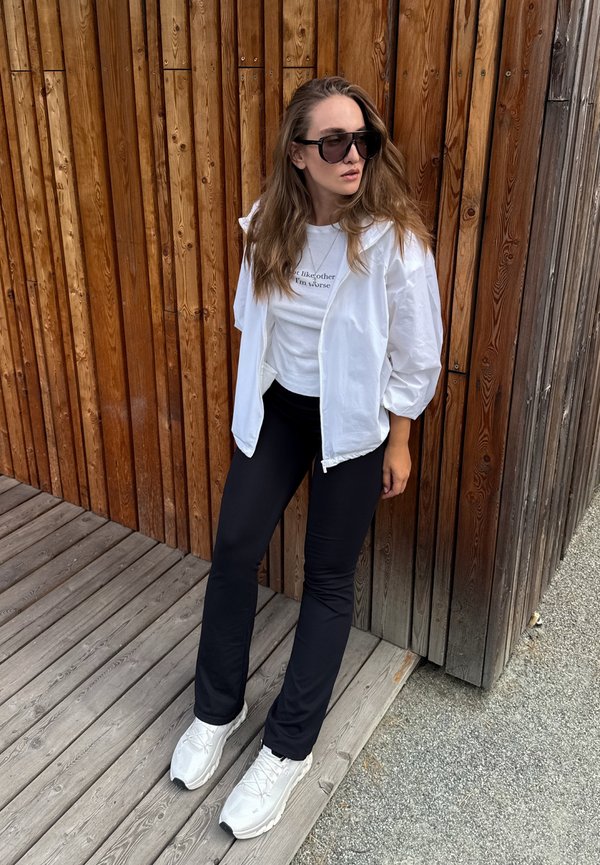 Vrouw met een wit jack, wit hemd, zwarte broek en witte sneakers, staand tegen een houten muur en kijkt naar links.