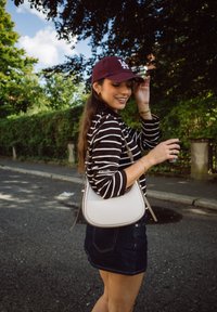 Borsa crossbody bianca con bordi arrotondati e dettagli beige; il modello indossa una maglietta a righe bianche e nere e una gonna di jeans, all'aperto su una strada soleggiata.