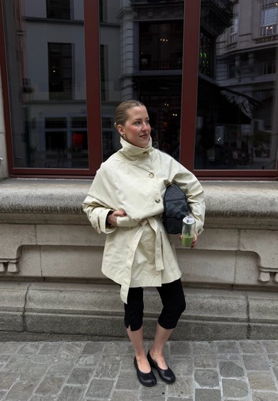 Mujer está de pie en la acera de la ciudad con un abrigo beige con cinturón, pantalones negros cortos y zapatos negros planos, sosteniendo una bebida verde y un bolso negro de hombro.