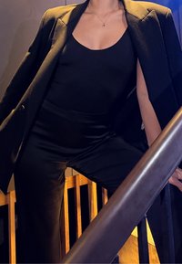 Persona che indossa un blazer nero appoggiato sulle spalle, una canotta nera senza maniche e pantaloni neri, in piedi vicino a una ringhiera di scale in legno.