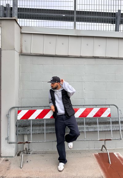 Chaleco negro sobre sudadera gris, pantalones oscuros y zapatillas blancas, de pie junto a una barricada de metal con franjas rojas y blancas contra una pared gris.