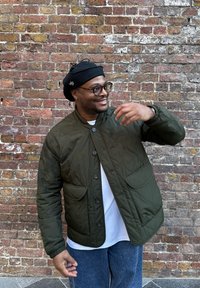 Blouson matelassé vert olive avec col montant, grandes poches frontales et fermetures à boutons, porté sur une chemise blanche et un jean bleu.