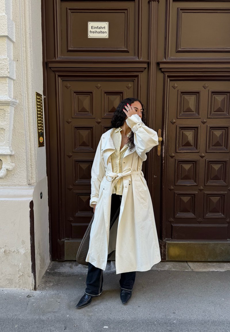 Femme en trench blanc et bottes noires debout, main près du visage, devant de grandes portes doubles marron avec un panneau en allemand.