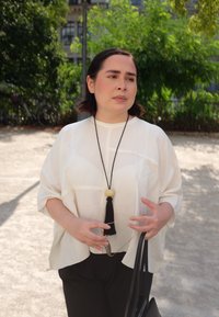 Blusa de lino blanca, holgada, con cuello redondo, collar de borlas negras y pantalones oscuros; sosteniendo gafas y un bolso negro.