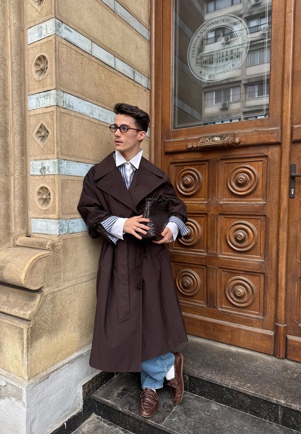 Junger Mann mit Brille, der einen langen dunklen Mantel, gestreiftes Hemd, blaue Jeans und braune Schuhe trägt, steht neben einer verzierten Holztür und hält eine schwarze Tasche.