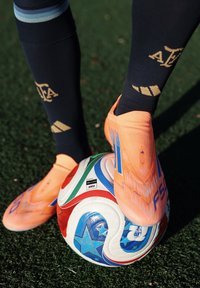 Joueur de football portant des chaussettes noires et des crampons orange vif contrôle un ballon de football multicolore sur un gazon vert.