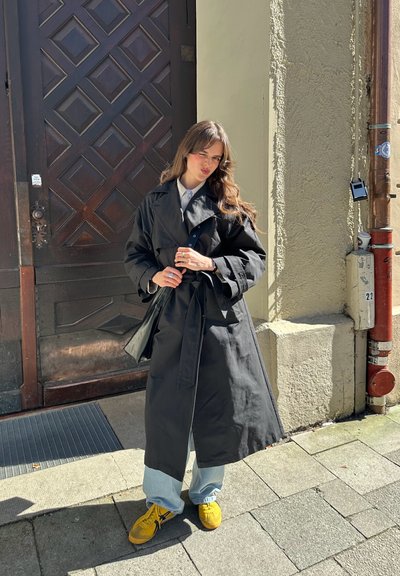 Abrigo negro de trench con cintura ceñida y cinturón, combinado con jeans claros y zapatos amarillos. El fondo presenta una puerta de madera y elementos urbanos.