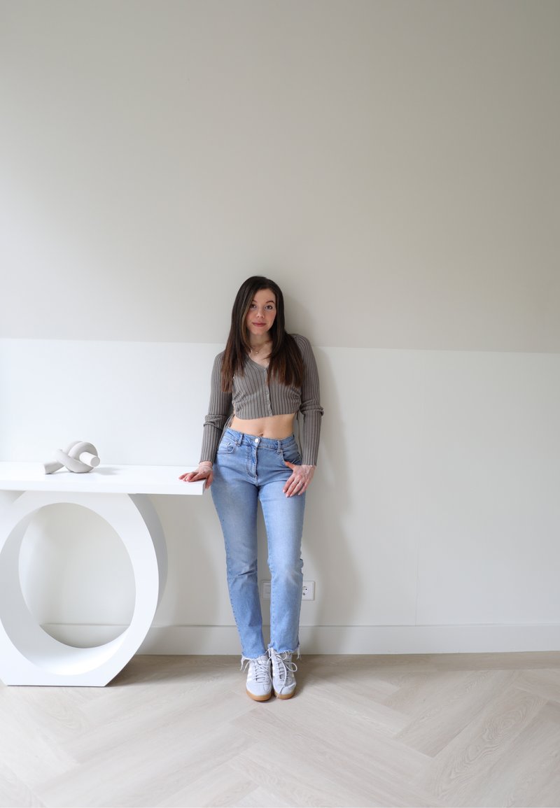 Γκρι ριμπεσμένος κοντός σπουδός, ανοιχτό μπλε τζιν με φθαρμένο τελείωμα και λευκά αθλητικά παπούτσια. Αφηρημένο λευκό γλυπτό πάνω σε ένα τραπέζι με στρογγυλά πόδια.