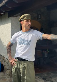 Bela majica s modrimi akcenti nosi napis "BROWN DUCKS", v kombinaciji s godrnastimi olivno zelenimi hlačami. Oseba nosi zeleni beret.