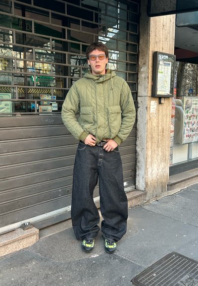 Joven vistiendo una chaqueta acolchada verde, vaqueros oscuros extragrandes, zapatillas de neón y gafas con lentes tintadas de naranja, de pie en una acera urbana.