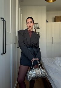 Blazer gris surdimensionné avec épaulettes, associé à une ceinture noire ajustée et une mini-jupe. Sac à main argenté en sequins avec des accents de nœud.