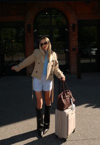 Beige trench coat, kék pulóver, csíkos rövidnadrág, fekete térdháig érő csizmák. Barna kézitáska és bézs bőrönd. Kültéri környezet, téglafal háttér.