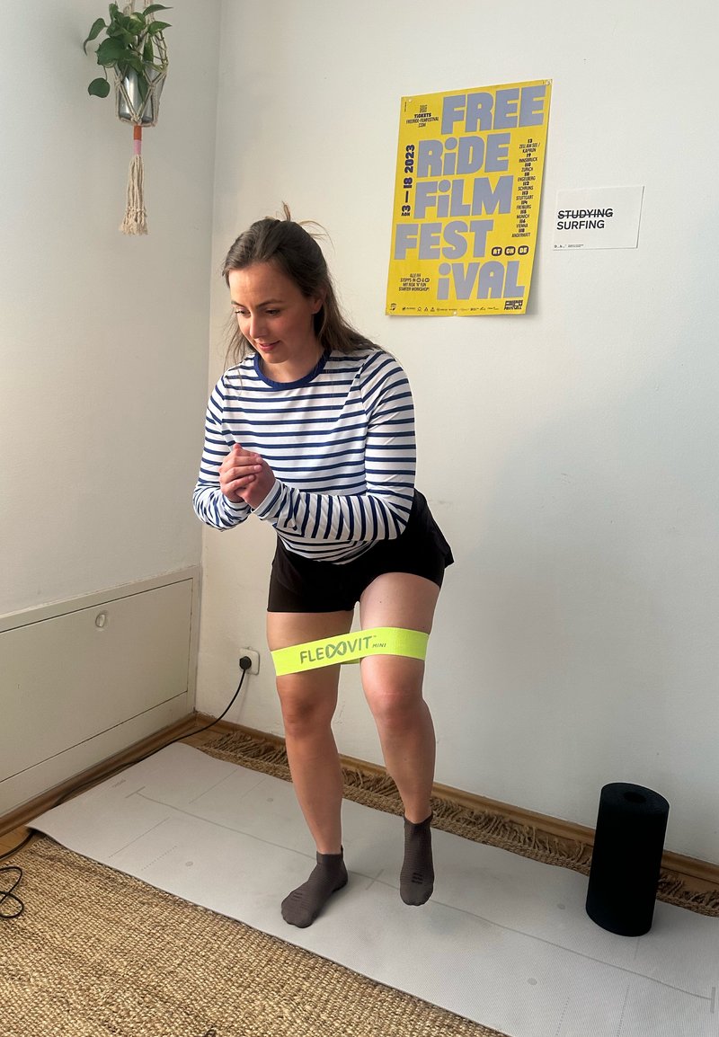 Femme faisant de l'exercice avec une bande de résistance jaune autour des cuisses sur un tapis de yoga dans une pièce avec des affiches au mur.
