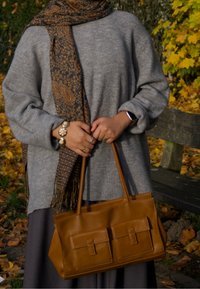 Sac à main en cuir marron avec deux poches avant, porté avec un pull gris et une écharpe à motifs, présentant une finition texturée et des poignées solides.