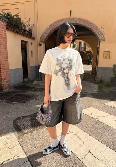 Persona con corte de cabello bob negro, que lleva una camiseta blanca gráfica de gran tamaño, pantalones cortos oscuros holgados, zapatillas slip-on a cuadros, sosteniendo una pequeña bolsa colorida.