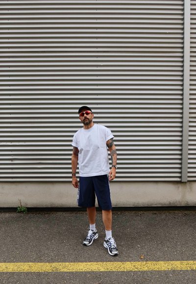 Camiseta blanca, pantalones cortos azul marino y zapatillas con acentos en negro y blanco. Tatuajes en los brazos, usando gafas de sol y una gorra negra. Fondo de pared texturizada.