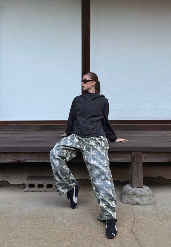 Femme portant une veste noire, des lunettes de soleil et un pantalon camouflage assise sur un banc en bois devant un mur blanc, regardant sur le côté.