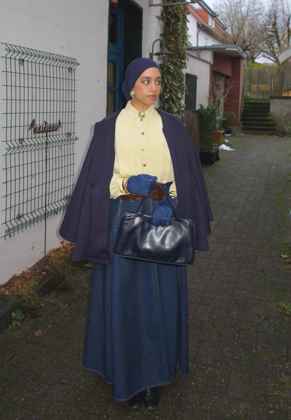 Donna con hijab blu navy, camicetta gialla, gonna di jeans e guanti che tiene una borsa nera in piedi in un vicolo all'aperto pavimentato.