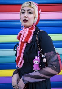 Strikket rødt og rosa skjerf og lue, lagt over en svart topp. Tilbehør inkluderer myke nøkkelringer i rosa og svart. Bakgrunnen har livlige striper.