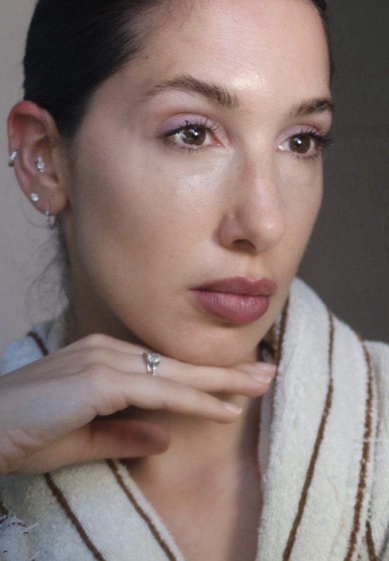 Femme avec une expression neutre reposant son menton sur sa main, portant un vêtement clair rayé et plusieurs piercings aux oreilles avec des anneaux et des clous.