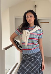 Pull rayé en bleu, rouge et vert. Associé à une jupe à carreaux grise. Tenant une pochette noire avec des ornements argentés. Pull blanc noué au cou.