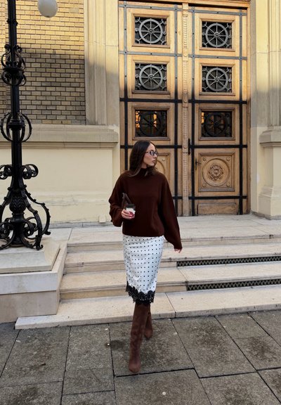 Mujer con suéter marrón, falda blanca con lunares y botas sostiene un café mientras baja unos escalones de piedra frente a grandes puertas de madera ornamentadas.