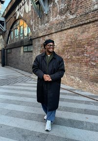 Manteau long noir, chemise verte, jeans bleus délavés, baskets grises. Debout sur un chemin pavé à motifs avec un mur en briques en arrière-plan.