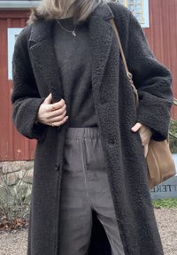 Manteau en laine long et texturé de couleur sombre avec un grand col, porté par-dessus un pull sombre et un pantalon en velours côtelé gris clair. Sac marron et accessoires visibles.