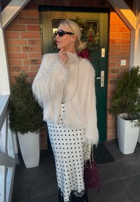 Femme aux cheveux blonds portant une veste légère en tissu duveteux, une jupe à pois, des lunettes de soleil et un collier de perles, debout près d'une porte verte ornée d'une couronne de Noël.