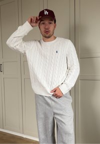 Jeune homme ajustant une casquette bordeaux LA, portant un pull blanc en maille torsadée et un pantalon de survêtement gris, debout à l'intérieur devant des armoires beiges.