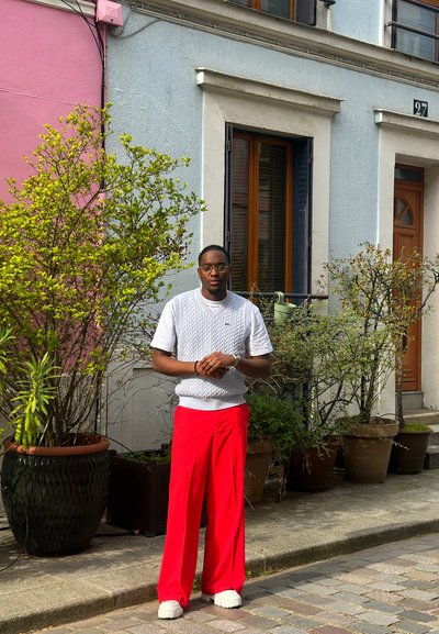 Hombre con chaleco de punto blanco y pantalones rojos de pie en una calle empedrada frente a edificios de colores pastel con plantas en macetas.