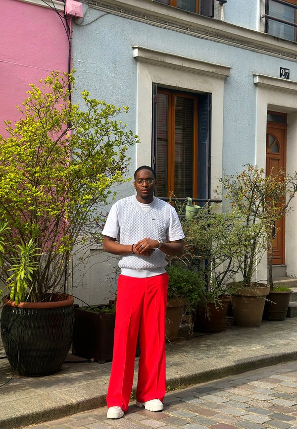 Mann iført hvit strikket vest og røde bukser står på en brosteinsgate foran pastellfargede bygninger med potteplanter.