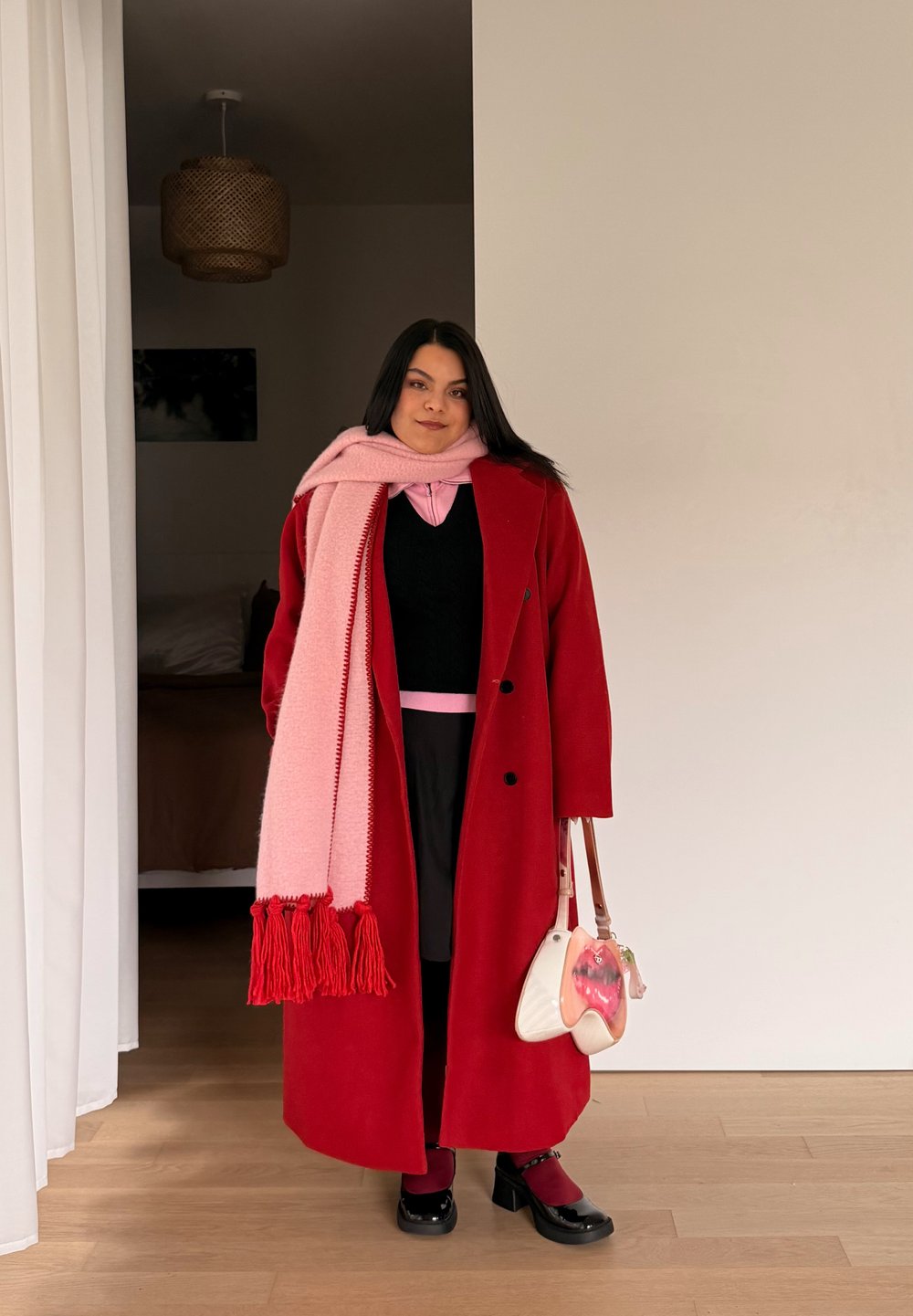 Femme portant un manteau rouge, une écharpe rose, une jupe noire et des chaussures noires, tenant un sac à main blanc et rose, debout à l'intérieur près d'un rideau blanc.