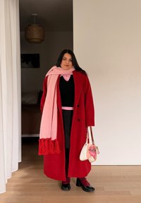 Femme portant un manteau rouge, une écharpe rose, une jupe noire et des chaussures noires, tenant un sac à main blanc et rose, debout à l'intérieur près d'un rideau blanc.