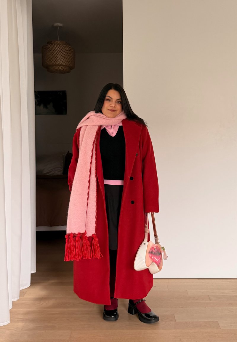Femme portant un manteau rouge, une écharpe rose, une jupe noire et des chaussures noires, tenant un sac à main blanc et rose, debout à l'intérieur près d'un rideau blanc.