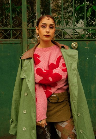 Mujer con gabardina verde sobre un suéter rosa y rojo, falda marrón y medias estampadas, de pie frente a una puerta metálica verde.