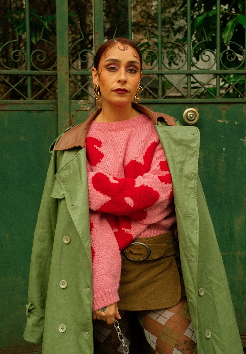 Femme portant un trench-coat vert sur un pull rose et rouge, une jupe marron et des collants à motifs, debout devant une porte en métal verte.
