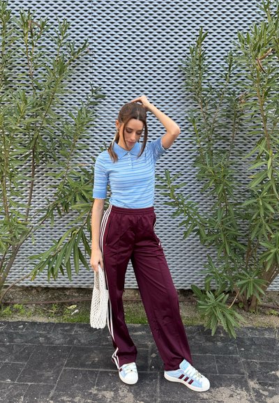 Mujer con polo de rayas azules y pantalones deportivos burdeos de pie sobre el pavimento, sosteniendo una bolsa blanca, frente a una pared texturizada con plantas verdes cerca.