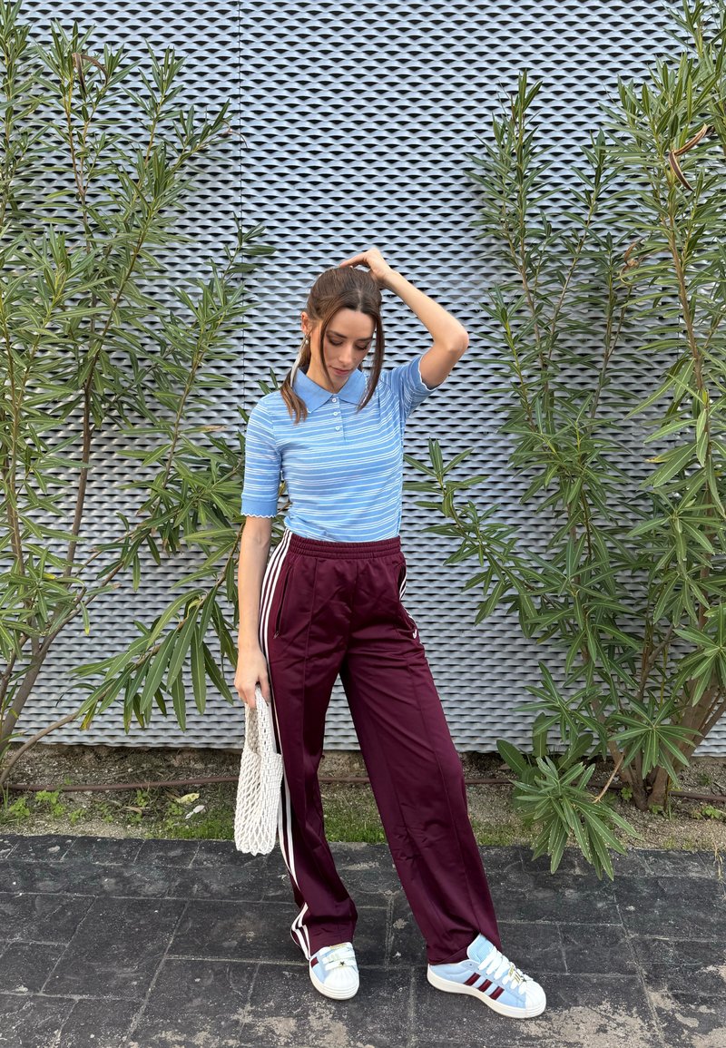 Kvinne i blåstripet polo og mørkerøde joggebukser står på fortauet, holder en hvit veske, mot en teksturert vegg med grønne planter i nærheten.