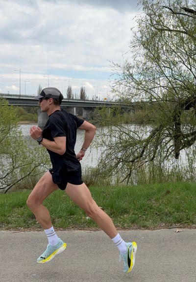 Hombre corredor con ropa deportiva negra y gorra corriendo por un sendero cerca del río con un puente y árboles de fondo en un día nublado.
