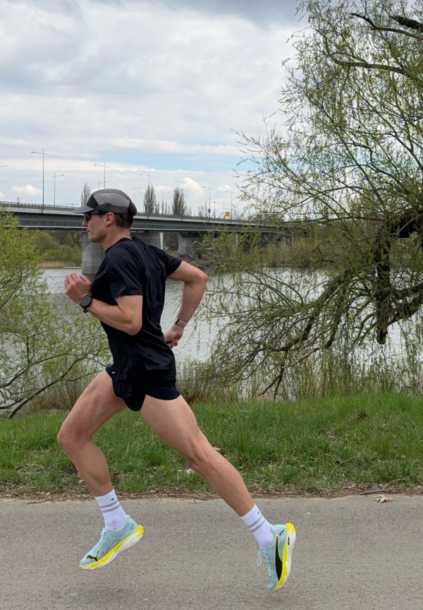 Muški trkač u crnoj sportskoj odjeći i kapi trči stazom blizu rijeke s mostom i drvećem u pozadini na oblačan dan.