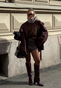 Bordeaux oversized jas, bruine trui, grijze minirok, kniehoge bruine laarzen. Model houdt een zwarte tas vast, draagt grote zonnebril en handschoenen.