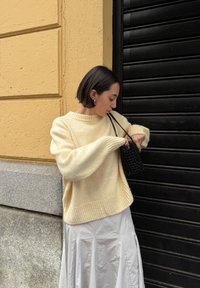 Pulôver de malha na cor creme com mangas oversized, combinado com uma saia plissada branca. Uma bolsa tecida preta adiciona contraste contra o fundo texturizado.